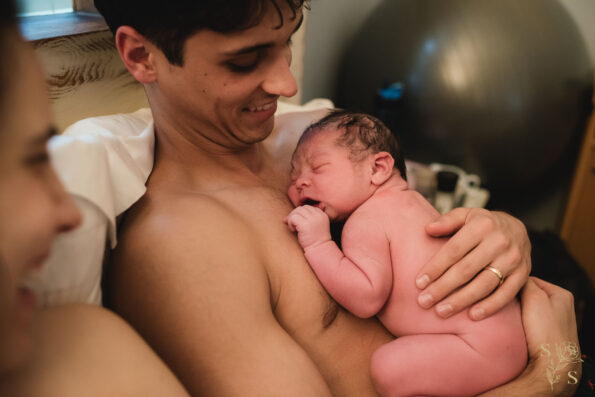 "Tender moment captured as a father holds his naked, newborn baby for the first time after a water birth, engaging in heartwarming skin-to-skin contact, creating a strong early connection."