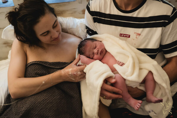 "Tender moment captured as an infant is cradled in the father's arms, wrapped in a towel, while the mother lovingly caresses the baby's face, expressing warmth and affection within the family."